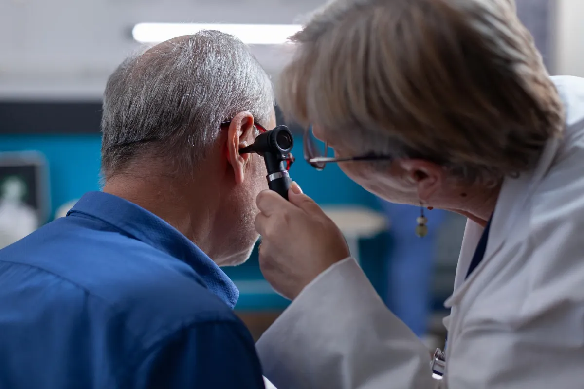 Woman doctor holding otoscope to do ear consultation for old man at medical appointment. Otologist using otology instrument to do examination and give medical advice to ill patient.